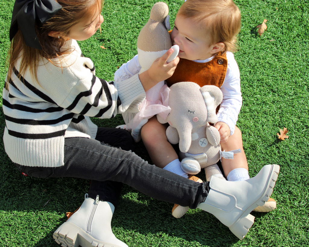 Little girl sitting on grass next to baby, playing with stuffed animals, wearing Trotters shoes on a bright sunny day.